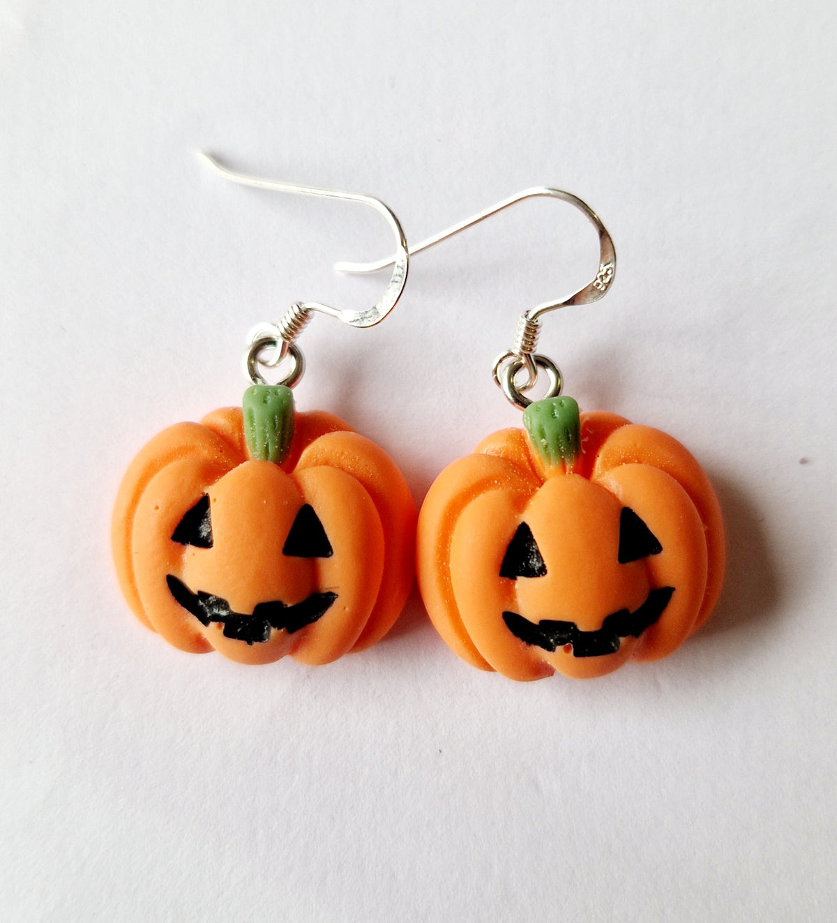 A pair of orange jack-o'-lantern dangle earrings with green stems and black facial features, suspended from sterling silver fishhook wires on a plain white background.