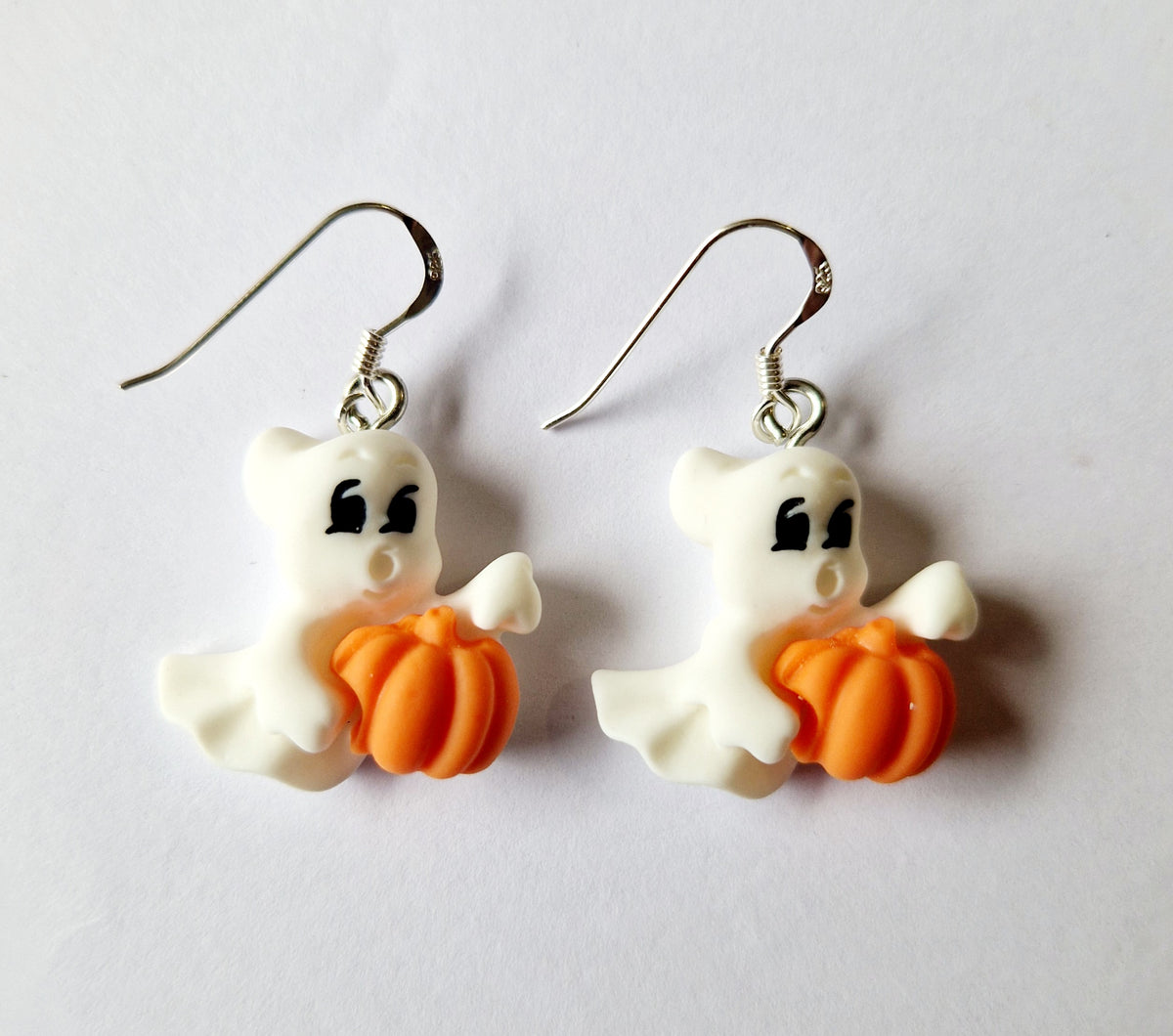 A pair of white ghost dangle earrings holding orange pumpkins, suspended from sterling silver fishhook wires on a plain white background.