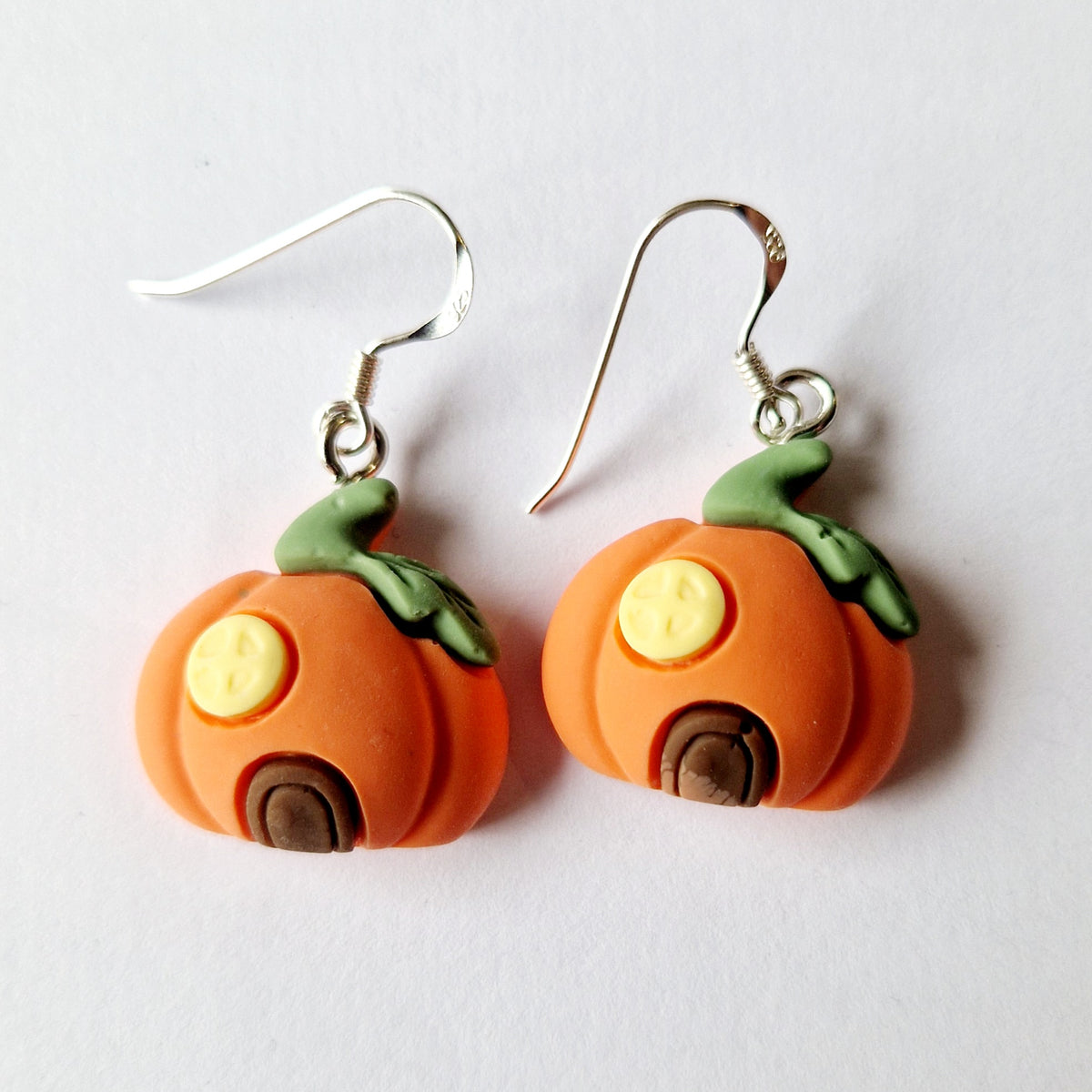 A pair of handmade pumpkin-shaped dangle earrings on a plain white background. The earrings feature orange pumpkin bodies with green stems, yellow button eyes, and brown door details, suspended from sterling silver fishhook wires.