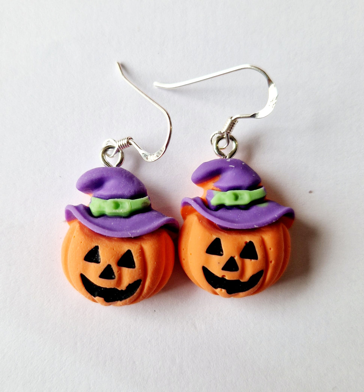 A pair of Halloween-themed dangle earrings featuring orange jack-o'-lantern pumpkins with carved faces, purple witch hats, and green bands, suspended from sterling silver fishhook wires on a white background.