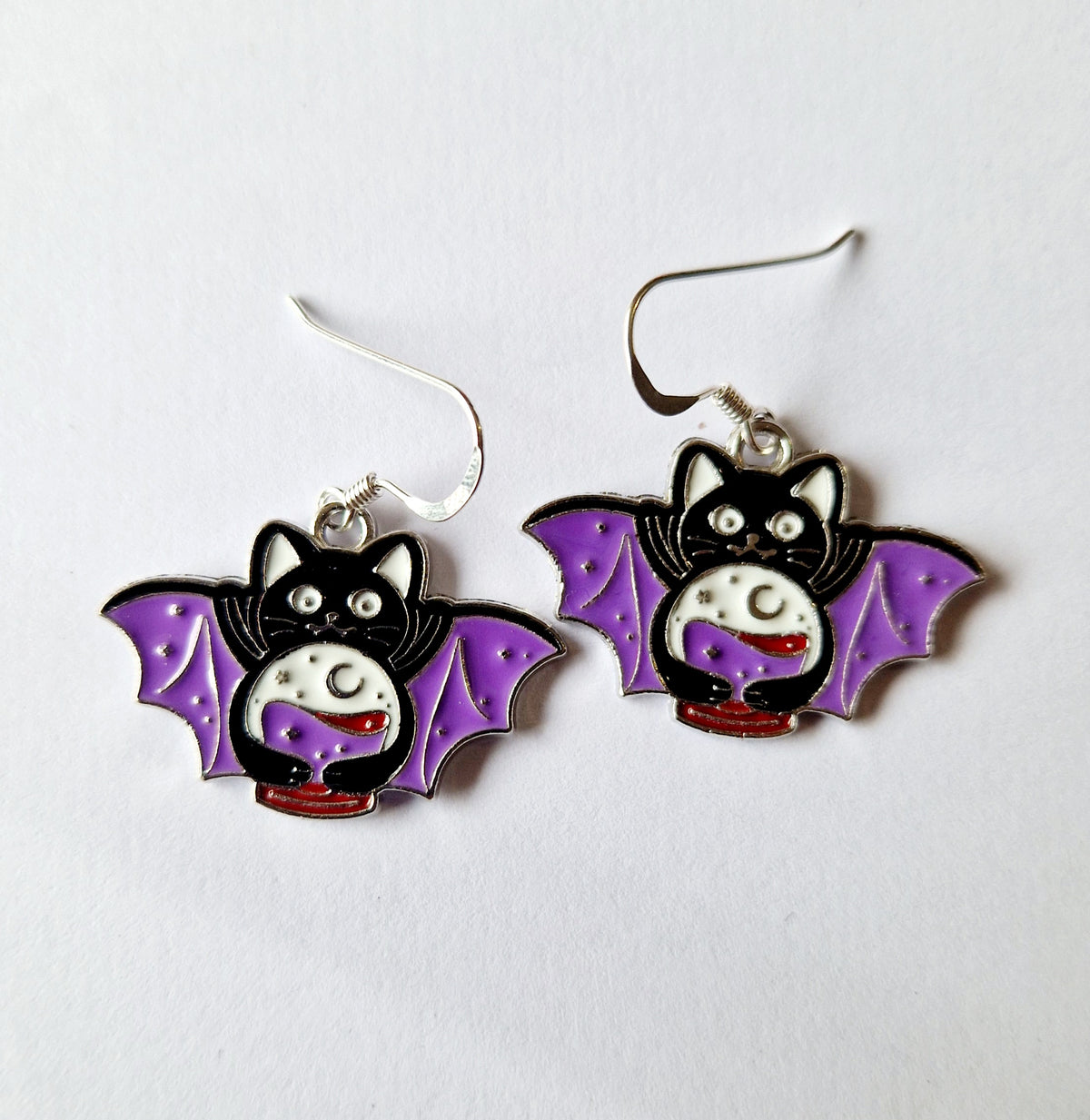 A pair of bat-shaped dangle earrings featuring a black bat body, purple wings, a white moon face, and a small red crescent moon on a plain white background.