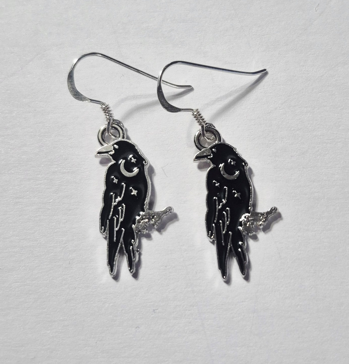 A pair of gothic-style raven drop earrings with black enamel bodies, sterling silver metal wire hooks, and small silver crescent moon and star details, displayed on a textured white surface.