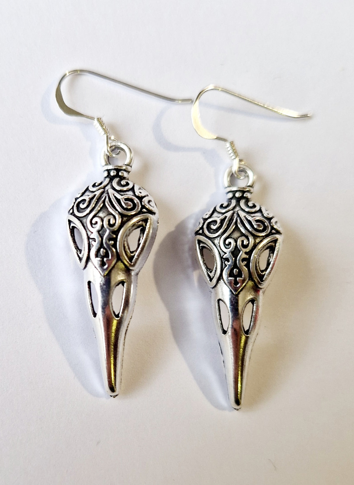 A pair of silver bird skull drop earrings with intricate filigree detailing and sterling silver fishhook wires, displayed on a plain off-white background.