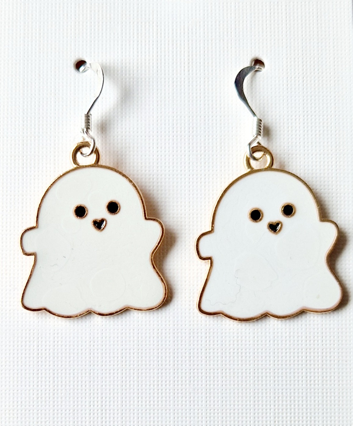 A pair of white ghost-shaped dangle earrings with gold-tone borders, sterling silver hooks, and cute black eyes and mouths, displayed on a textured white background.