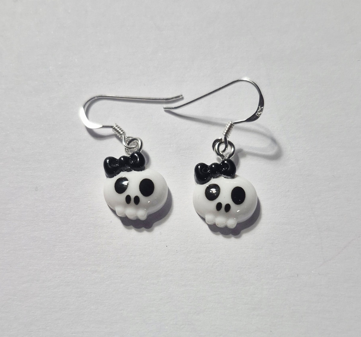 A pair of dangling skull earrings with white skull charms, black bow details, and sterling silver fishhook wires, displayed on a light gray textured background.