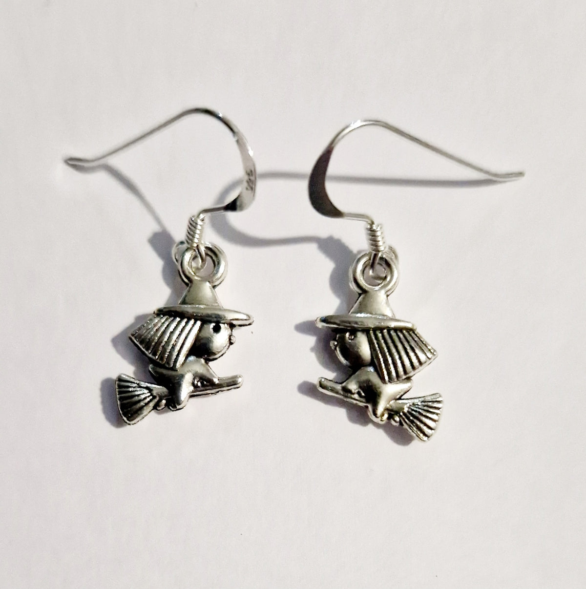 A pair of silver-toned dangle earrings featuring cartoon witch charms riding broomsticks, shown against a plain white background.