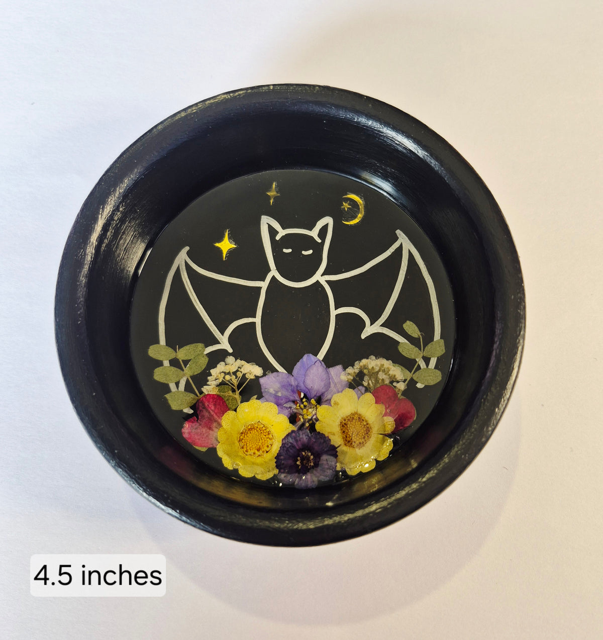 A round black ceramic bowl with a hand painted gothic bat design, featuring a silver bat outline, Golden stars and moon, and pressed flowers at the bottom. The text '4.5 inches