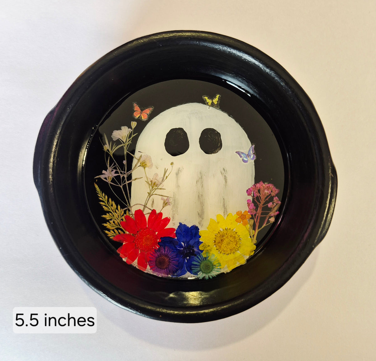 A round black decorative bowl featuring a hand painted white ghost design with dried flowers and butterflies inside, shown on a plain light background. A small label in the lower left reads 5.5inches