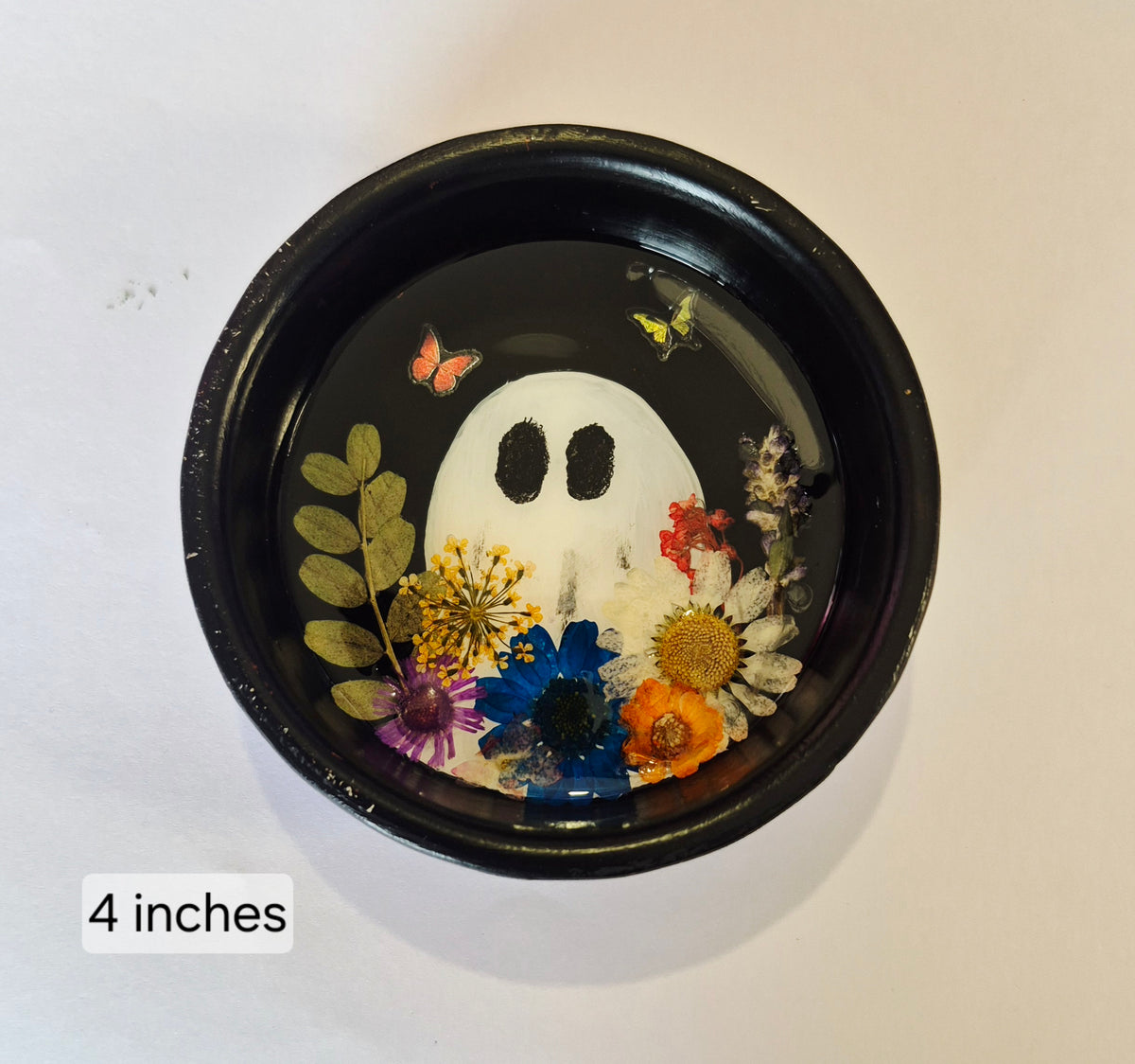A round black decorative bowl containing a hand painted white ghost design surrounded by pressed flowers and butterflies under clear resin, shown on a white background with a '4 inches' size label.