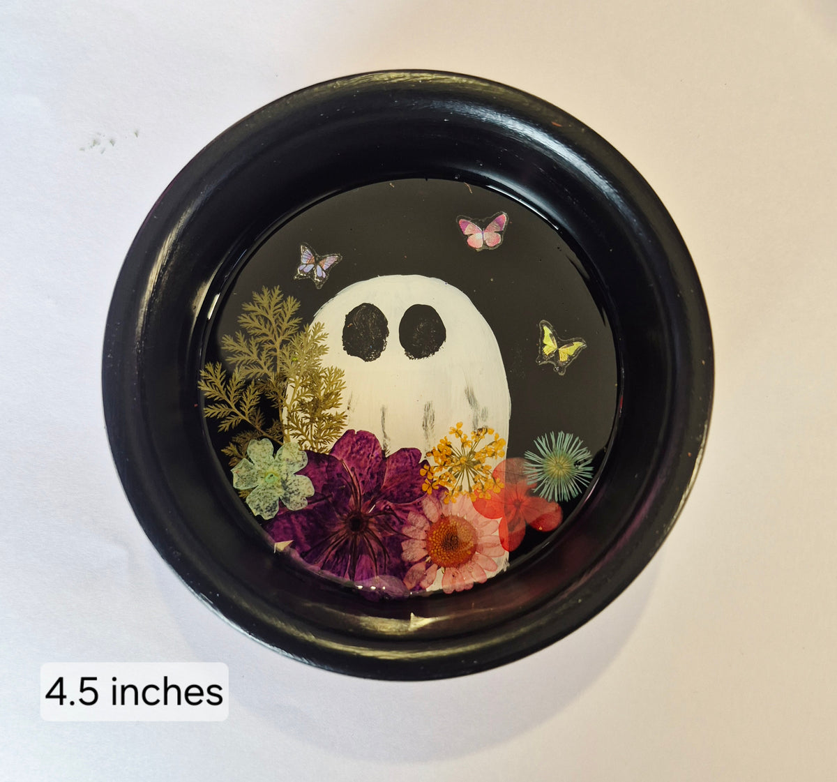 A round black tray with a hand painted white ghost face surrounded by colourful pressed flowers and butterflies on a resin background, shown on a white surface with a '4.5 inches' size label in the lower-left corner.