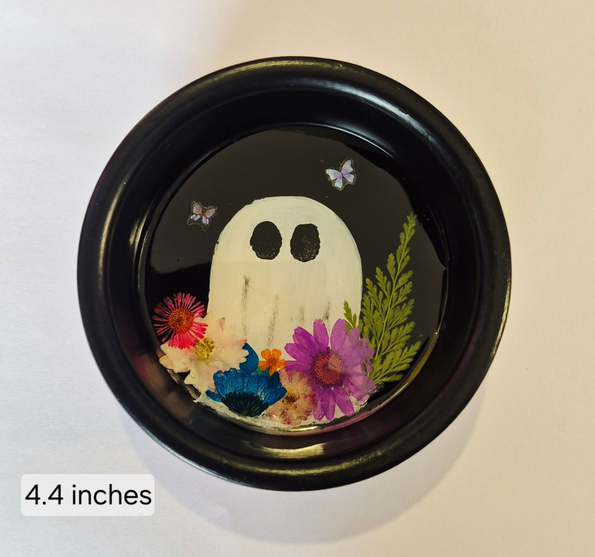A round black tray containing a hand painted white ghost face surrounded by colourful pressed flowers and green leaves, photographed from above on a light background. A small label in the lower left reads '4.4 inches