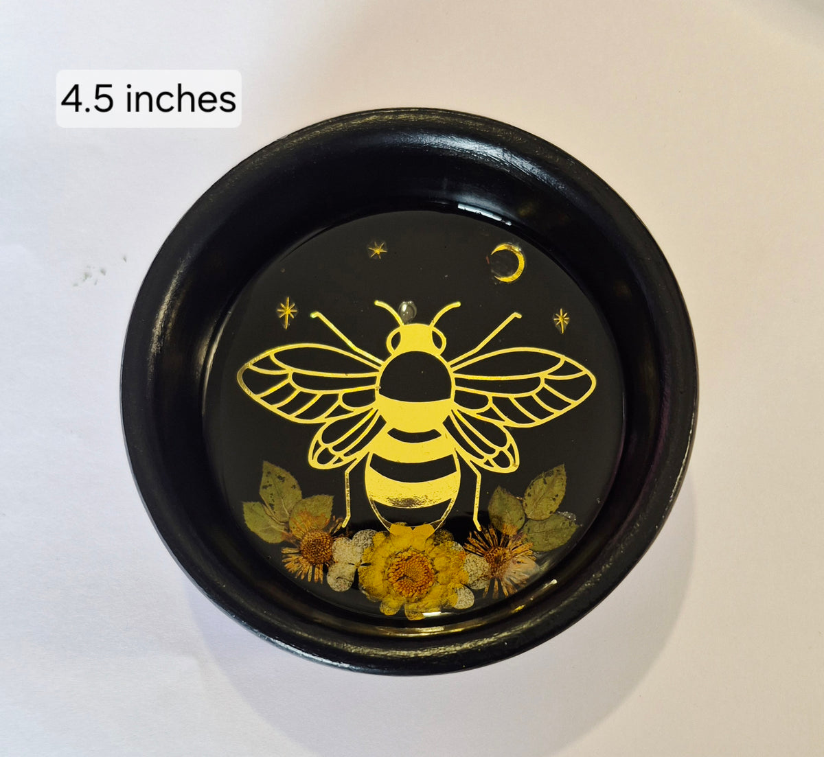 A round black decorative dish or bowl with a resin finish, featuring a gold bee design, small yellow flowers, and dried botanicals inside. The text '4.5 inches