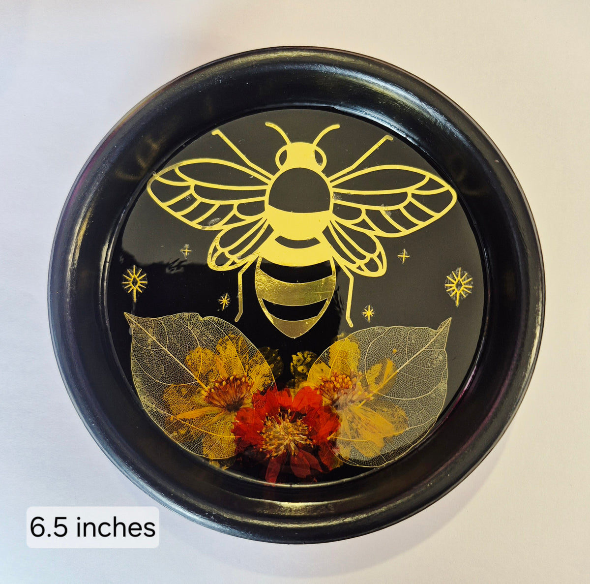 A round black decorative tray or dish with a resin finish, featuring a gold bee design above yellow and red pressed flowers and translucent leaves. The text '6.5 inches