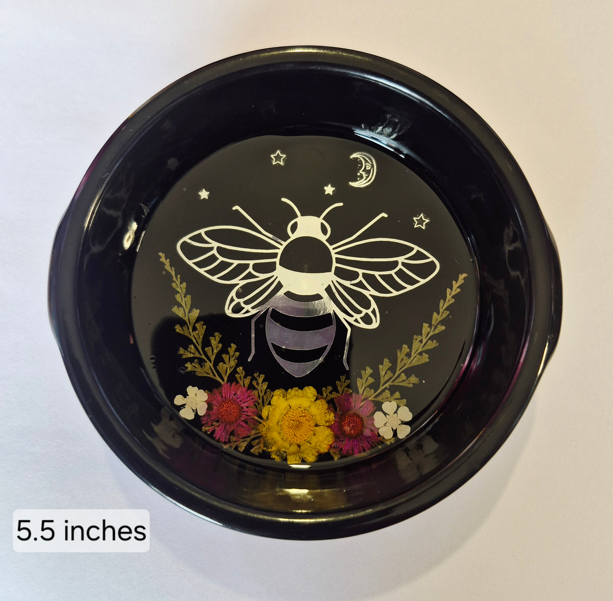 A round black decorative bowl or dish with a resin finish, featuring a bee design, surrounded by small pressed flowers in yellow, pink, and white. The bowl is photographed from above on a plain light background, with a text label in the lower left that reads '5.5 inches
