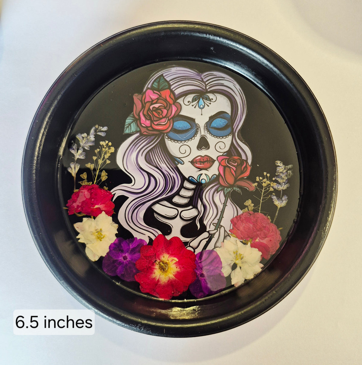 A round black decorative tray featuring a Day of the Dead sugar skull illustration with pressed flowers, shown on a plain light background. A text overlay in the lower left reads '6.5 inches