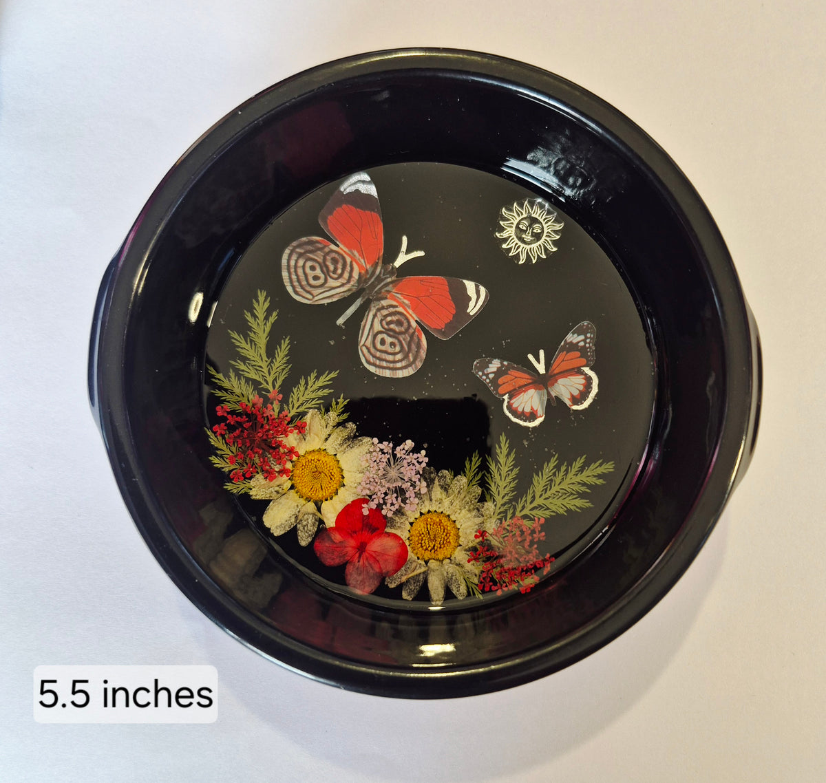 A round black decorative bowl with a resin finish, featuring a design of butterflies and pressed flowers on the inside. The text '5.5 inches