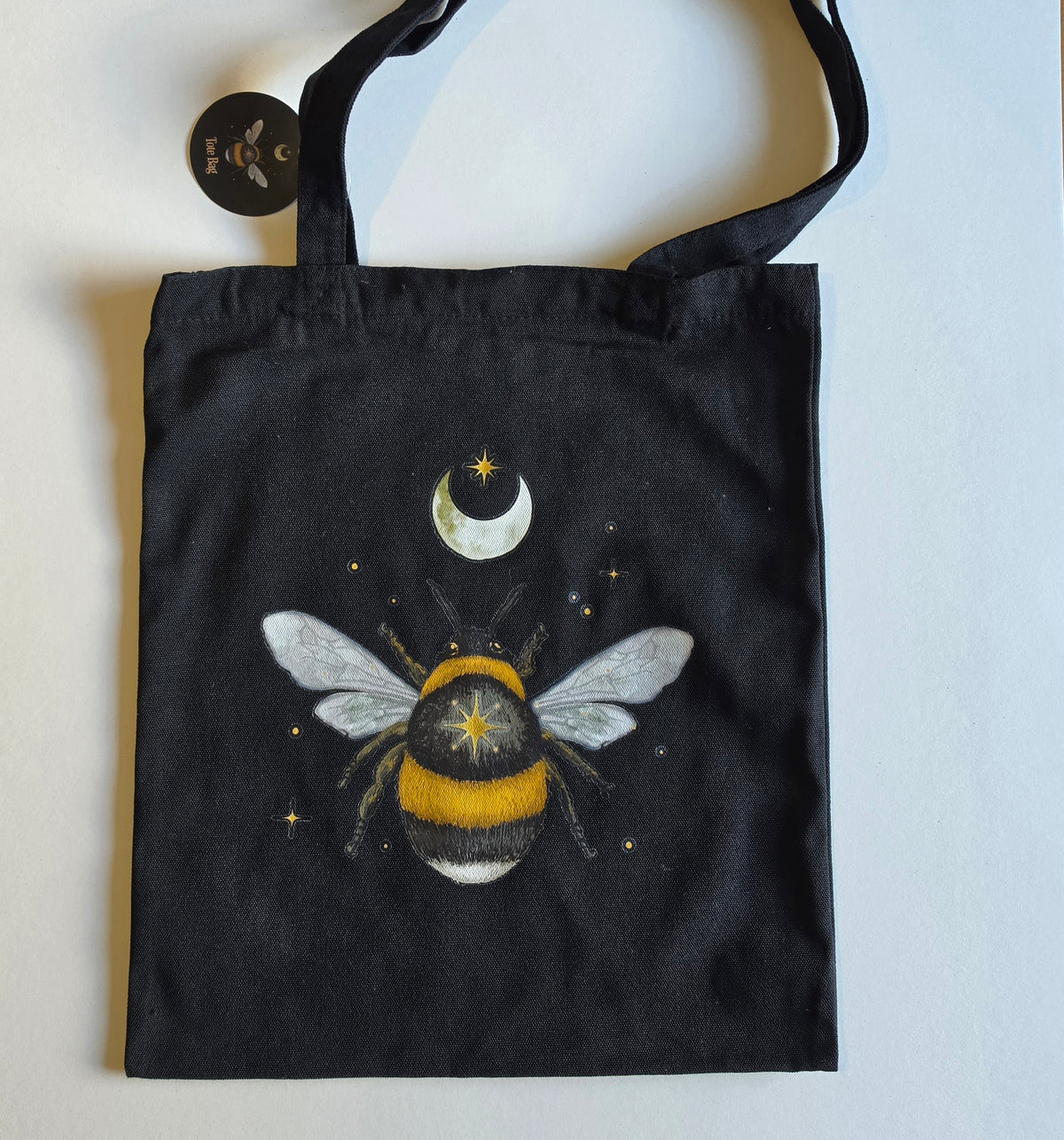A black canvas tote bag featuring a whimsical illustration of a bumblebee, crescent moon, and stars on a light gray background.