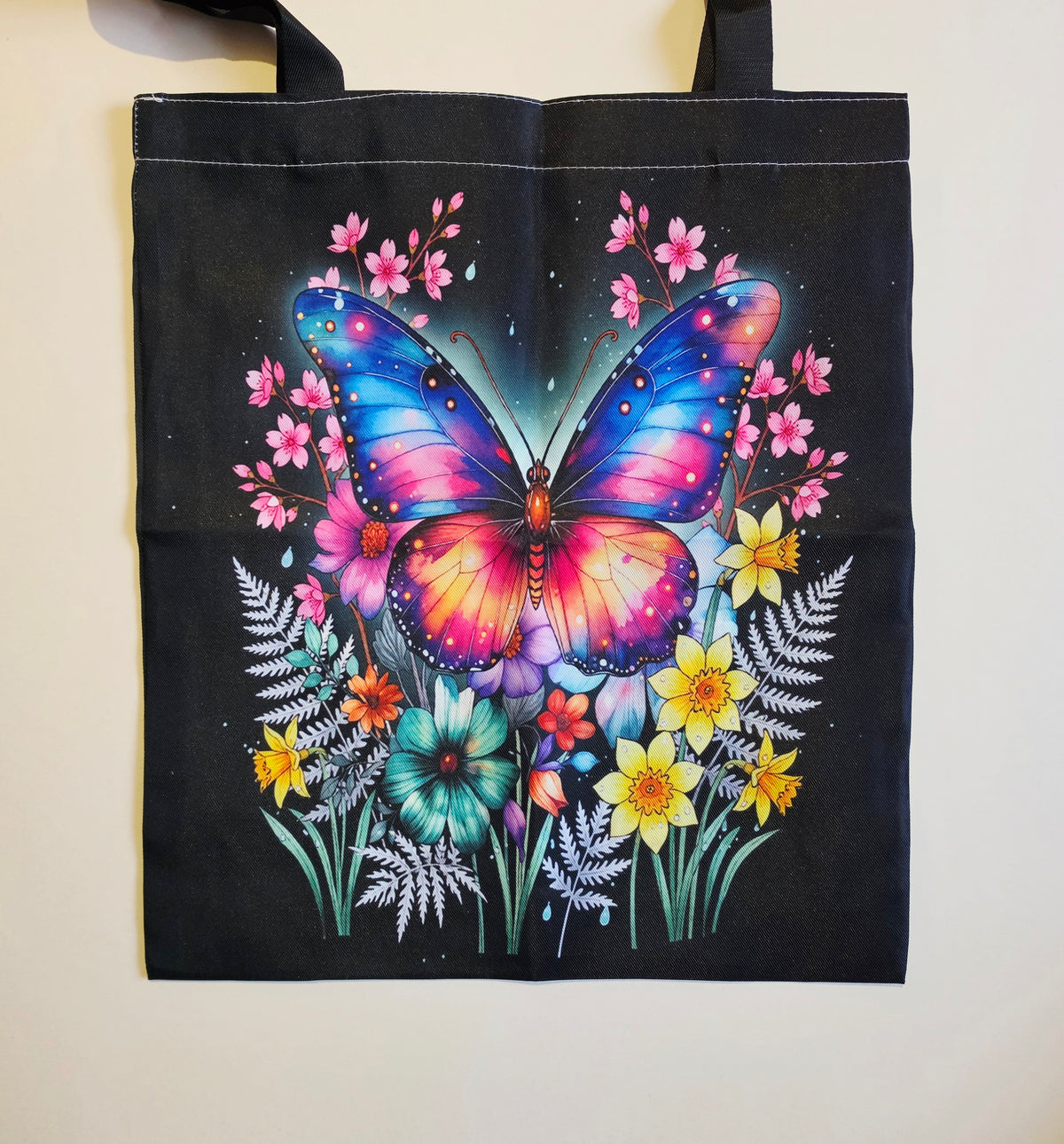 A black tote bag featuring a vibrant, colorful graphic of a butterfly surrounded by flowers, hanging against a plain off-white wall.