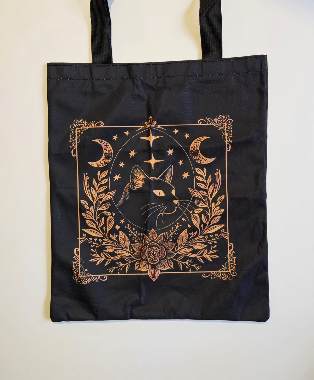 A black tote bag with black handles, featuring a central graphic print of a black cat surrounded by gold celestial and floral motifs, displayed against a plain off-white background.