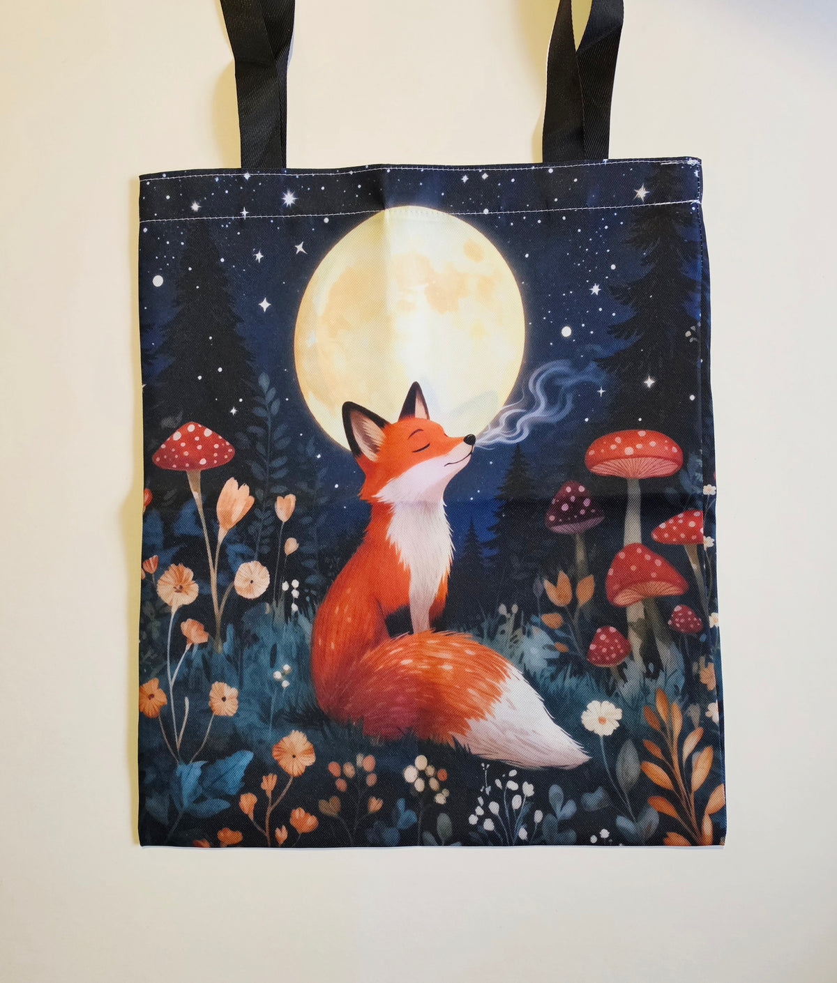 A fabric tote bag with black shoulder straps, featuring a printed illustration of an orange fox sitting under a large full moon in a starry night sky, surrounded by red and white mushrooms and wildflowers.