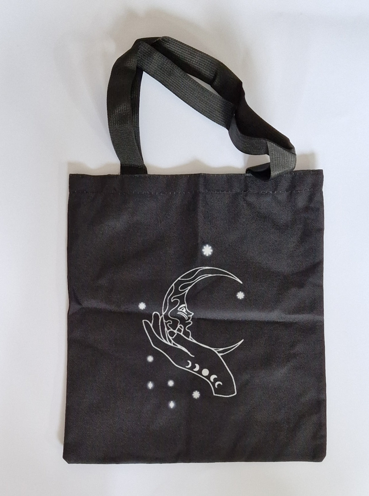 A black canvas tote bag with black shoulder straps, featuring a white line art graphic of a crescent moon, a hand, and stars on the front, set against a plain white background.