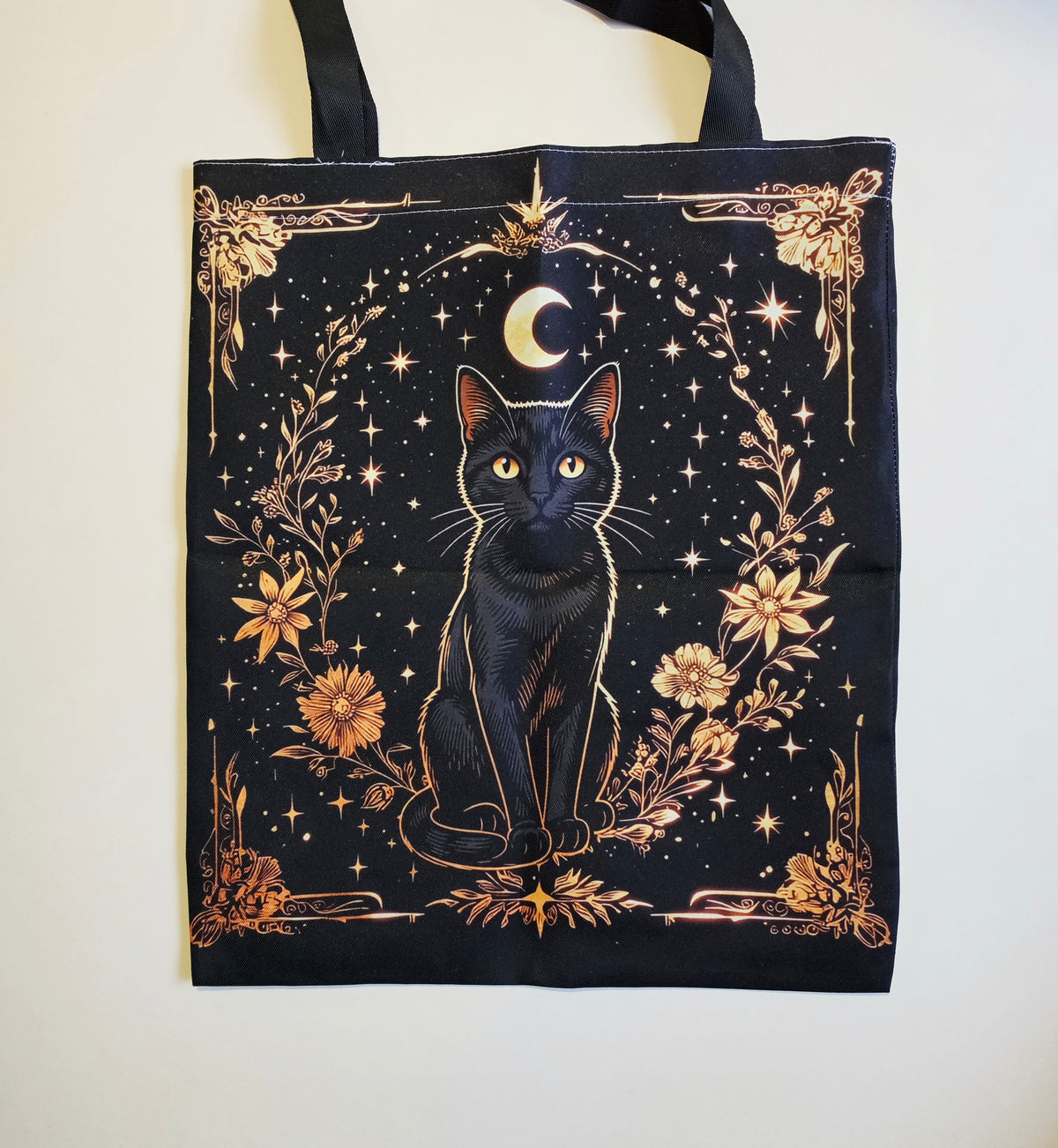 A black tote bag featuring a detailed graphic print of a black cat under a crescent moon, surrounded by gold-toned stars, flowers, and decorative borders, hanging against a plain off-white wall.