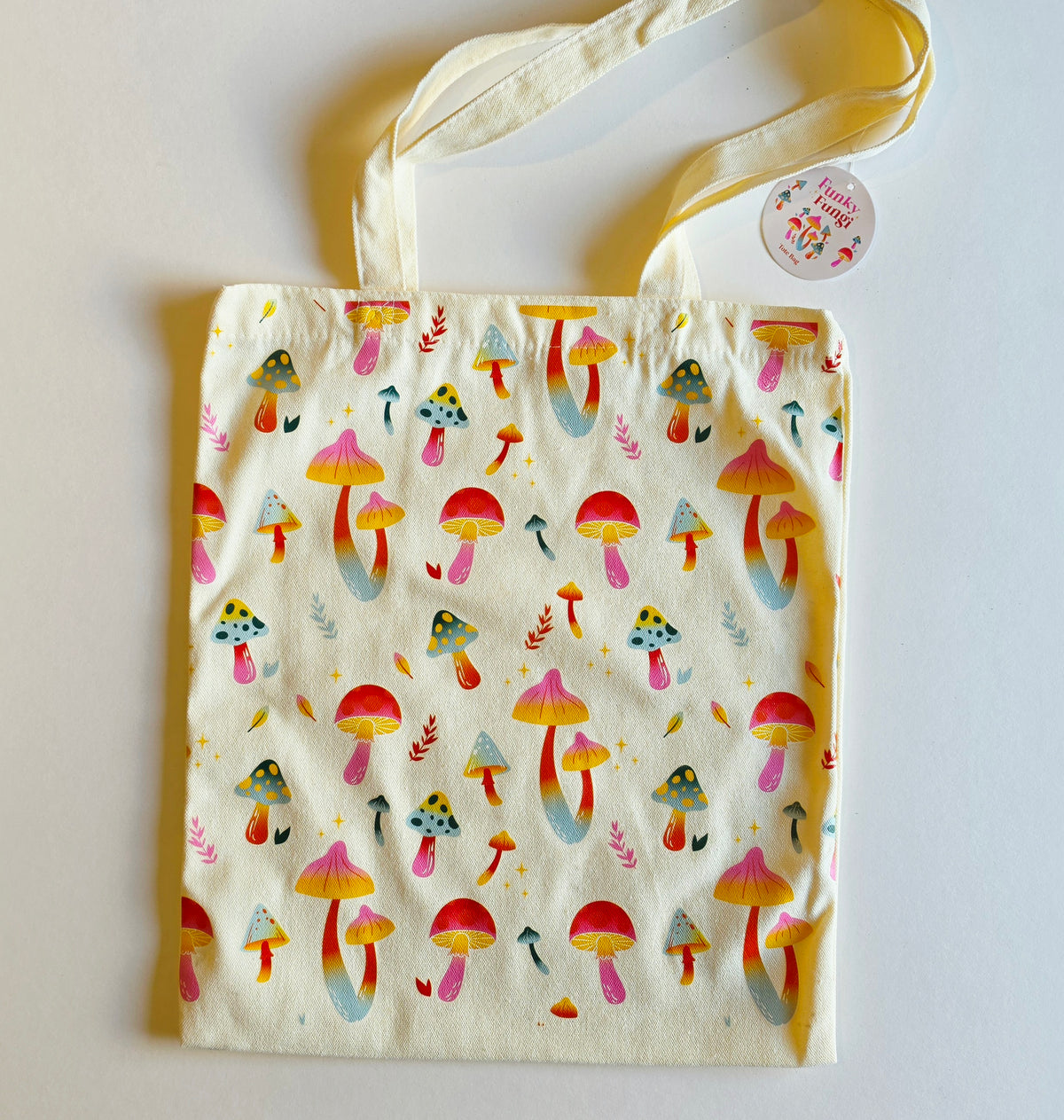 A cream canvas tote bag with a colorful mushroom print, featuring two shoulder straps and a round paper brand tag on the right side.