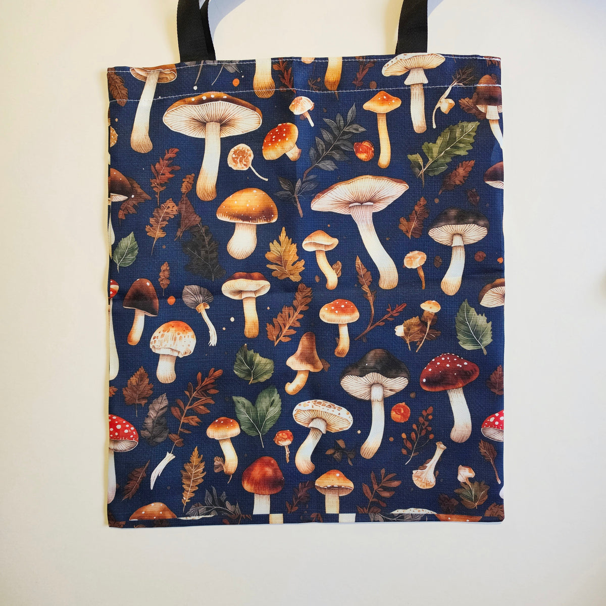 A fabric tote bag with a dark navy blue background and an all-over print of illustrated mushrooms, leaves, and small berries. The bag has black shoulder straps and is photographed hanging against a plain off-white wall.