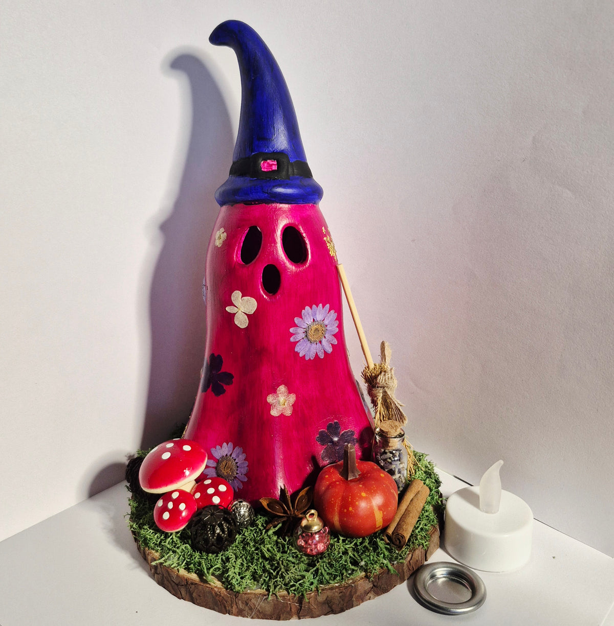A handmade whimsical Halloween witch ghost LED candle holder, Cone burner shaped like a pink ghost with a blue pointed hat, decorated with flowers, mushrooms, a small pumpkin, and a tiny figure, displayed on a faux moss base with a tea light candle nearby.
