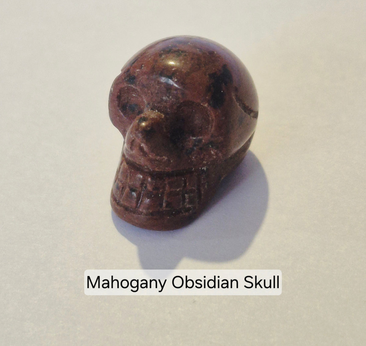 A carved Mahogany Obsidian skull figurine on a light beige background with a text label overlay reading 'Mahogany Obsidian Skull'.