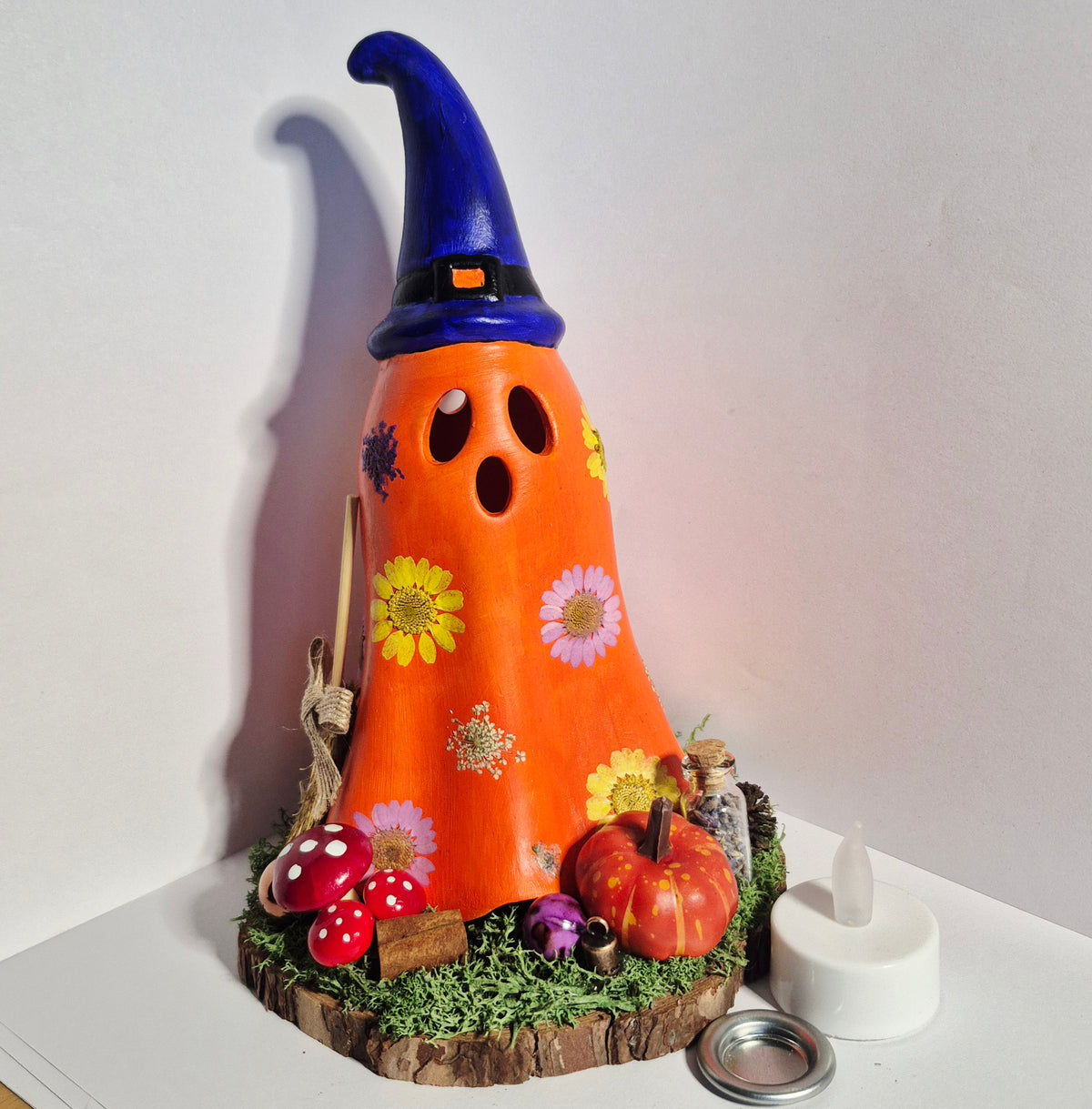 A handmade orange ghost-shaped Halloween decoration with a blue witch hat, decorated with flowers, set on a mossy wooden base with small decorative props, shown on a white surface against a white wall.