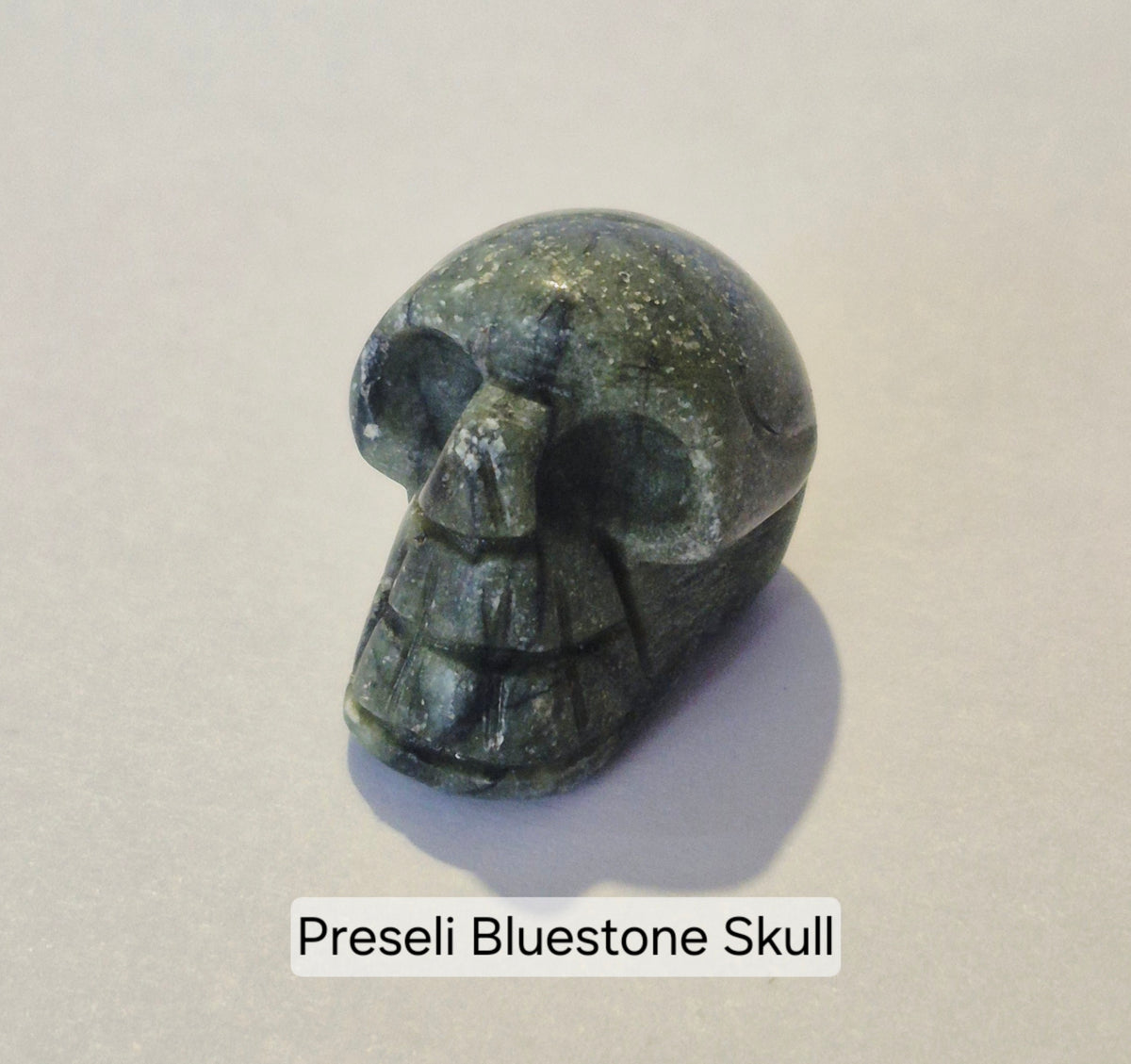 A carved Preseli bluestone skull charm or bead, showing natural stone texture in muted greenish-gray and olive tones, photographed on a plain light background with a text label underneath.