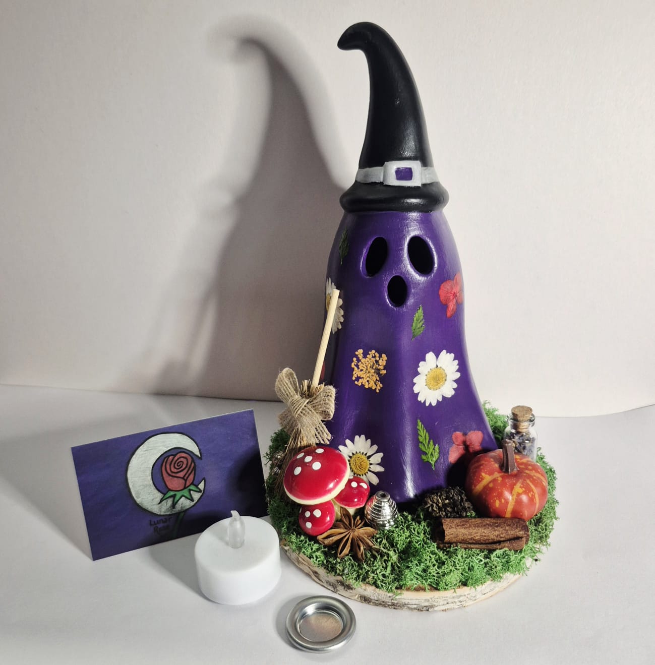 A purple decorative Witchy Ghost shaped cone burners on a white surface against a plain wall, with real pressed flowers and magical trinkets with a small LED candle, cone tray and card beside it.