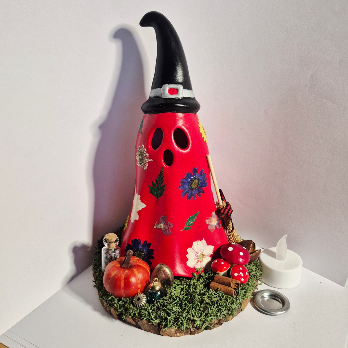 A handmade decorative Ghost candle holder with a red hood, black pointed hat, and autumn-themed embellishments including a small pumpkin, mushrooms, moss, and a broom, displayed on a white surface against a plain wall.