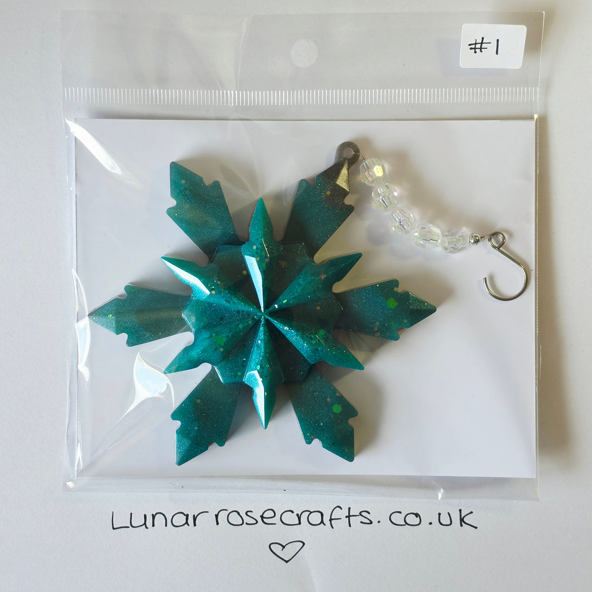 A teal glittery snowflake-shaped hangie with a clear beaded chain and silver hook clasp, packaged in a clear plastic bag on a white backing card. A handwritten label says '#1' and the card shows lunarrosecrafts.co.uk