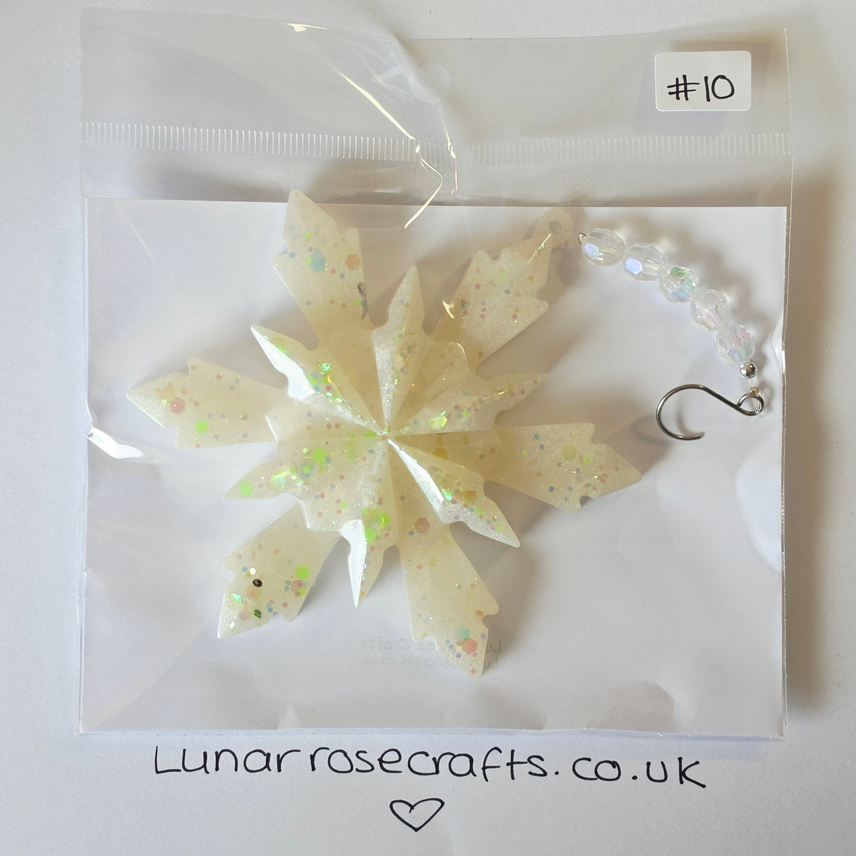 A cream glittery snowflake-shaped hangie with a clear beaded chain and silver hook clasp, packaged in a clear plastic bag on a white backing card. A handwritten label says '#10' and the card shows lunarrosecrafts.co.uk