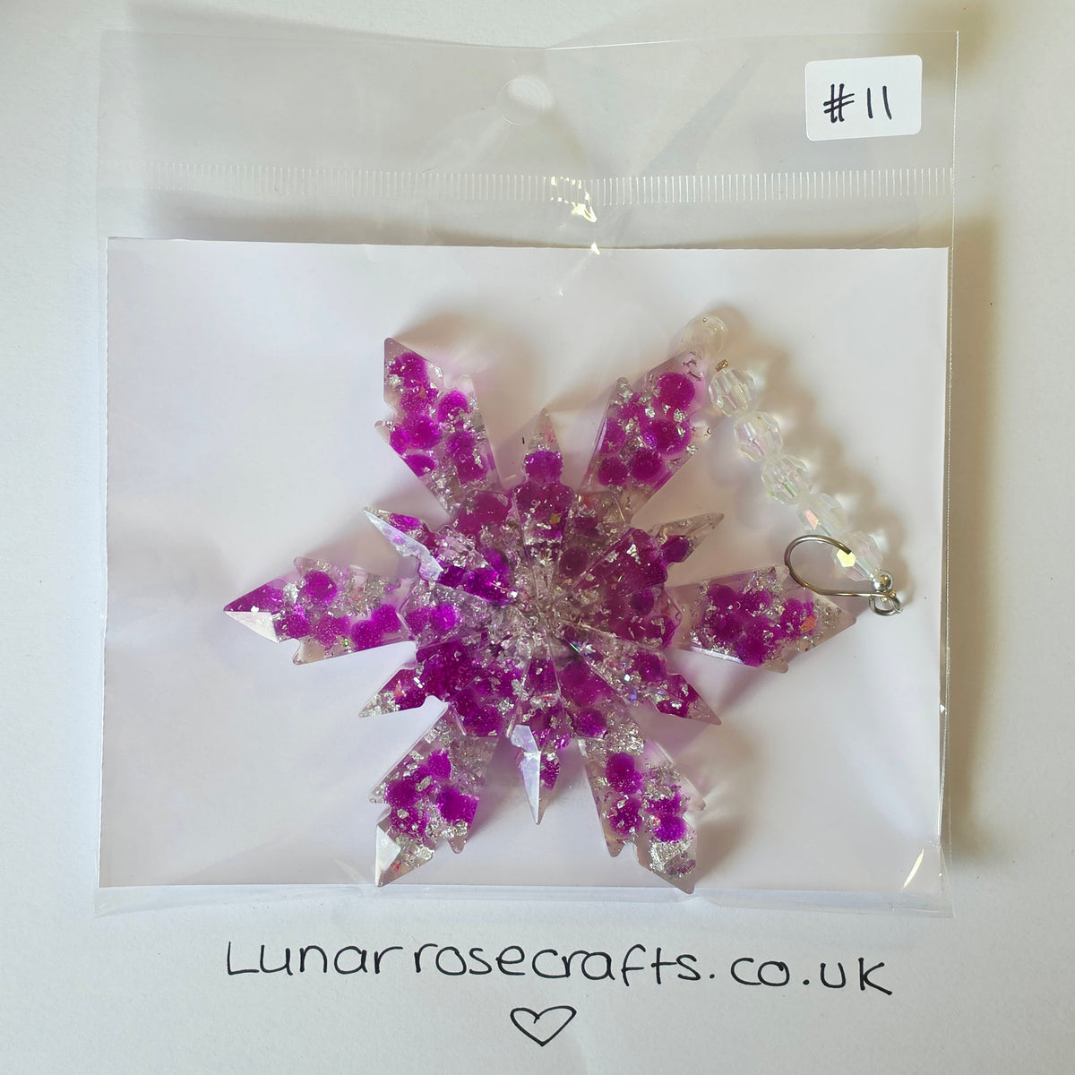 A snowflake-shaped resin ornament with embedded purple crystals and silver foil, packaged in a clear plastic bag on a white backing card. A handwritten label reads '#11' and the card is branded 'Lunarrosecrafts.co.uk'