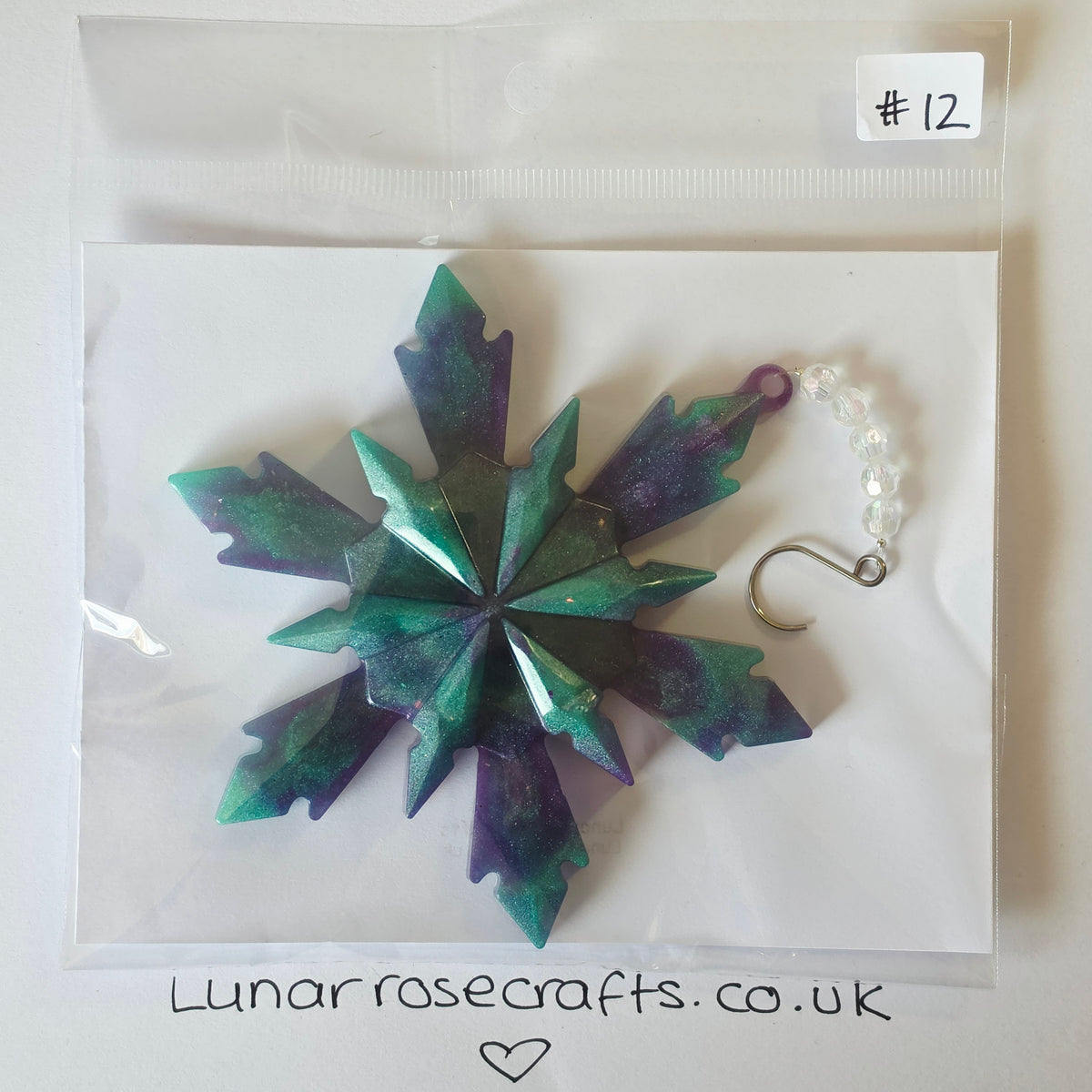 A green and purple glittery snowflake-shaped hangie with a clear beaded chain and silver hook clasp, packaged in a clear plastic bag on a white backing card. A handwritten label says '#12' and the card shows lunarrosecrafts.co.uk