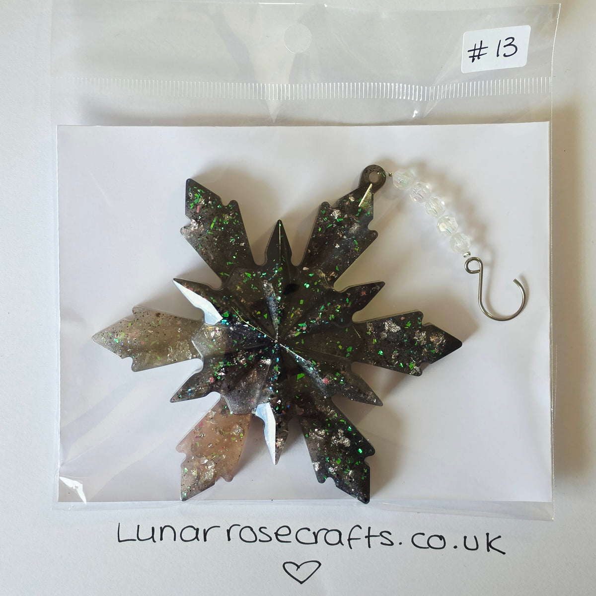 A packaged snowflake-shaped hangie with a dark glittery center and translucent outer points, attached to a white beaded loop and metal hook, shown inside a clear plastic bag on a white backing card. A handwritten label says '#13' and the card shows 'Lunarrosecrafts.co.uk'