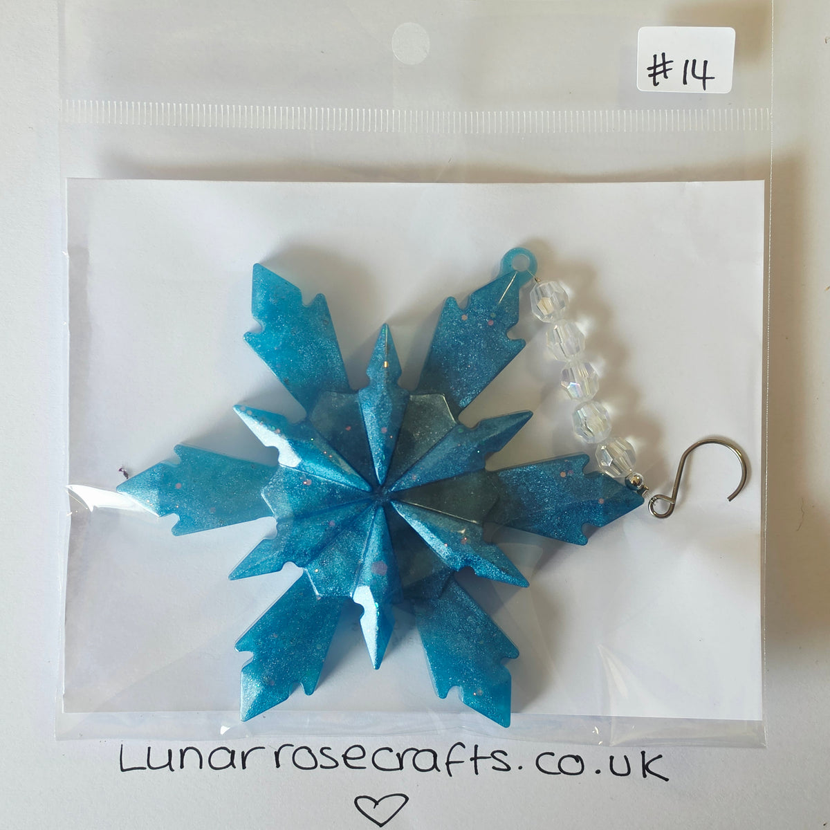 A blue glittery snowflake-shaped hangie with a silver hook and clear bead chain, packaged on a white card inside a clear plastic bag. Handwritten text at the bottom reads 'Lunarrosecrafts.co.uk'