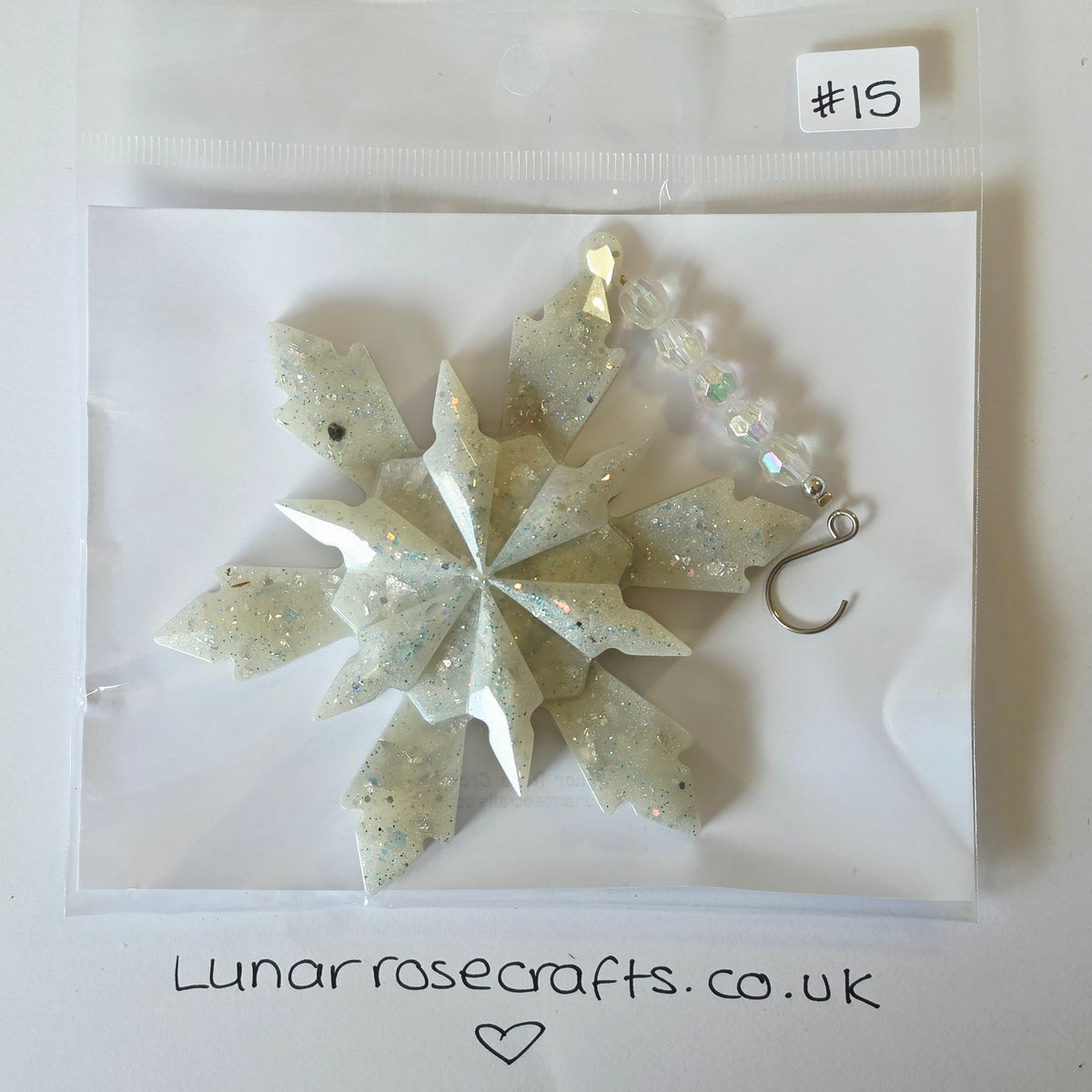 A glittery white snowflake ornament with iridescent accents, packaged in a clear plastic bag on a white backing card. A handwritten sticker reading '#15' is visible in the top right, and 'Lunarrosecrafts.co.uk'