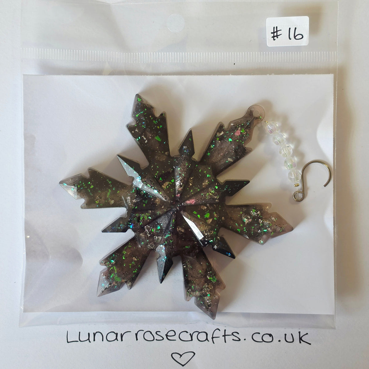 A faceted snowflake-shaped resin ornament with embedded glitter, sealed in a clear plastic bag with a handwritten label '#16' and the website Lunarrosecrafts.co.uk