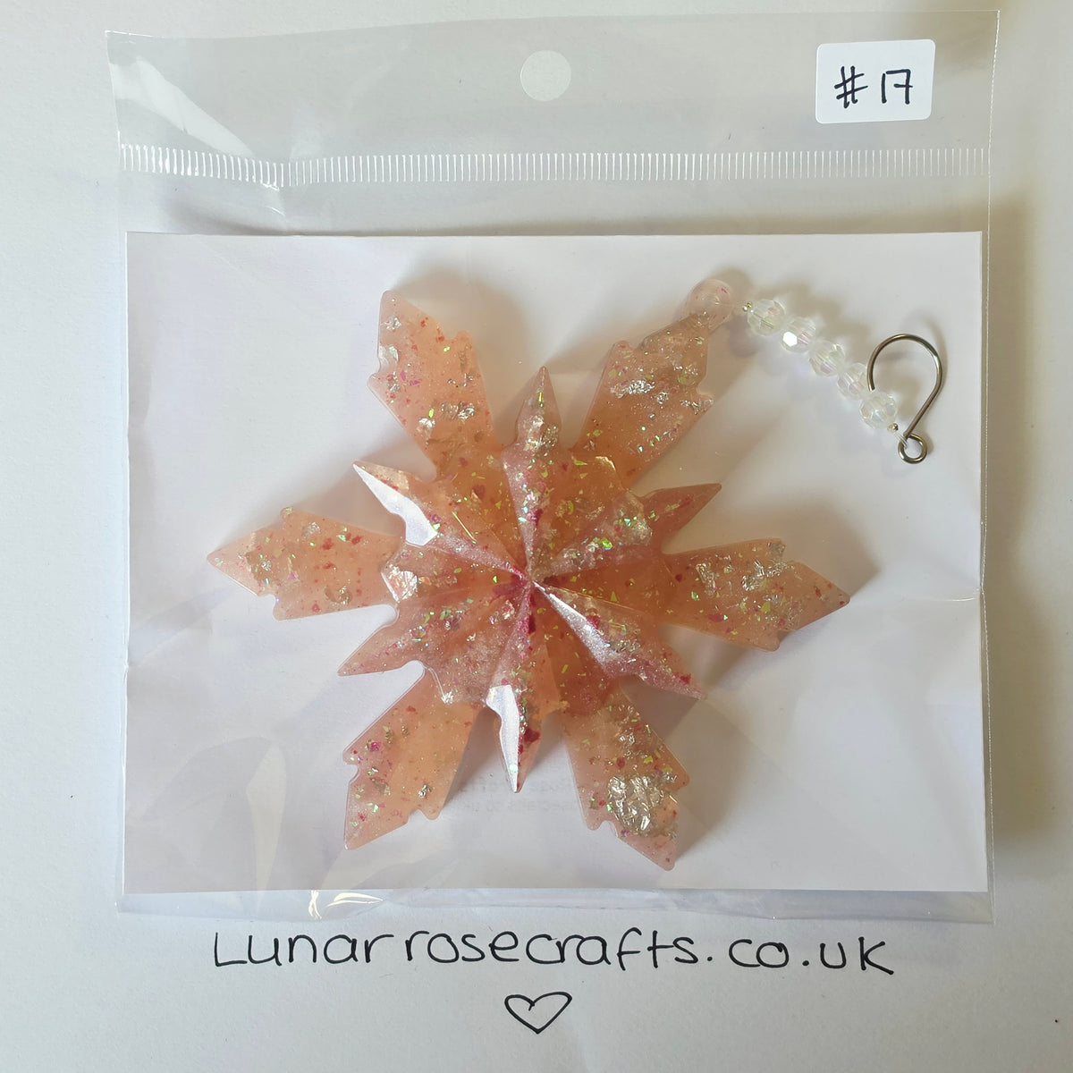 A translucent peach-pink resin snowflake ornament with embedded glitter, displayed on a white backing card inside a clear plastic bag. A white tag at the top right reads '#17', and the card is branded with 'Lunarrosecrafts.co.uk'