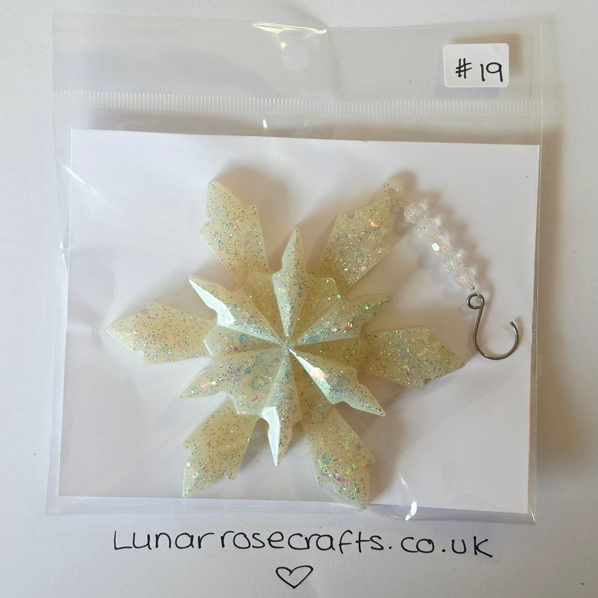 A glittery snowflake ornament in a clear plastic bag with a white card backing. The ornament has iridescent glitter and a small beaded hook charm.