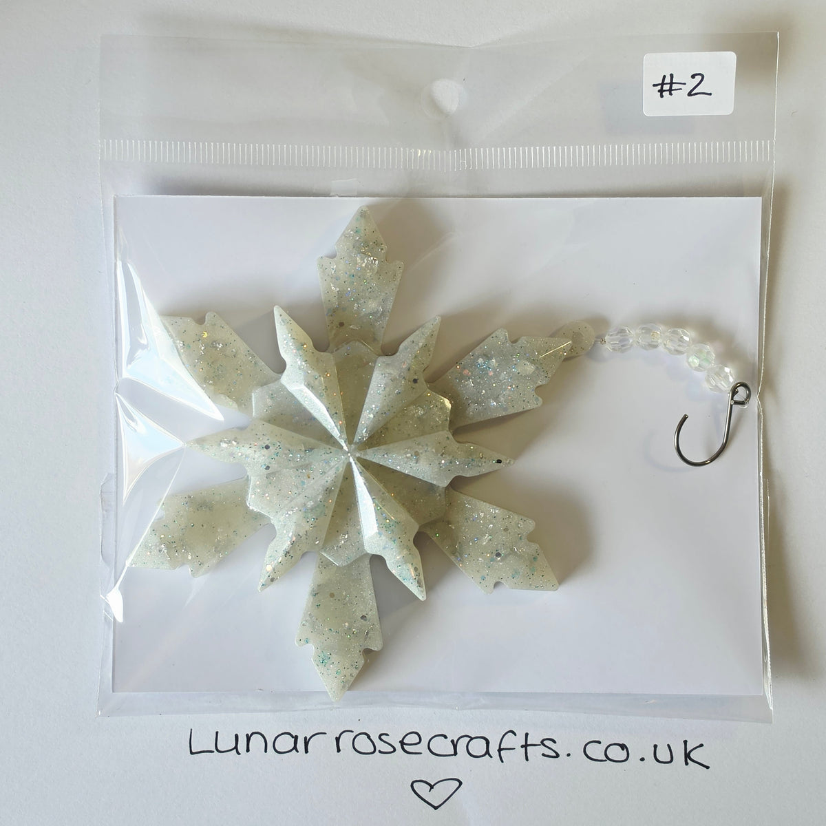 A handmade snowflake-shaped hangie by Lunar Rose Crafts, packaged in a clear plastic bag with a white backing card. made of textured, glittery off-white resin with silver and blue flecks, and has a small silver hook attached on the right side.