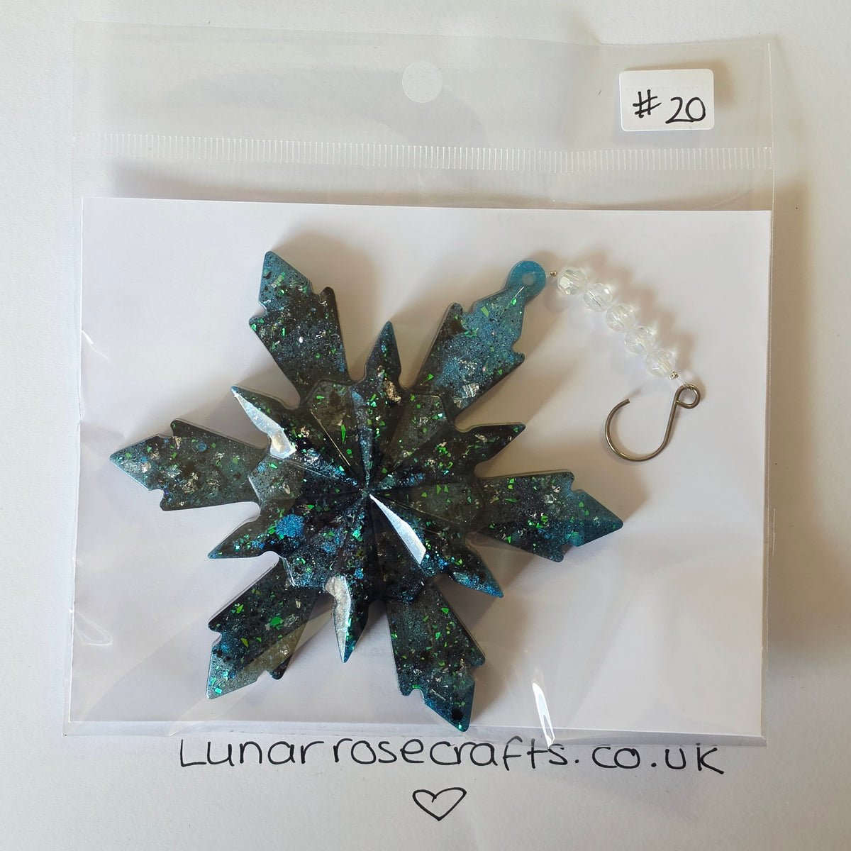 A glittery snowflake-shaped hanging ornament in dark teal and green with holographic sparkle, packaged on a white card inside a clear plastic bag. A small white tag reads '#20', and the card shows the website Lunarrosecrafts.co.uk