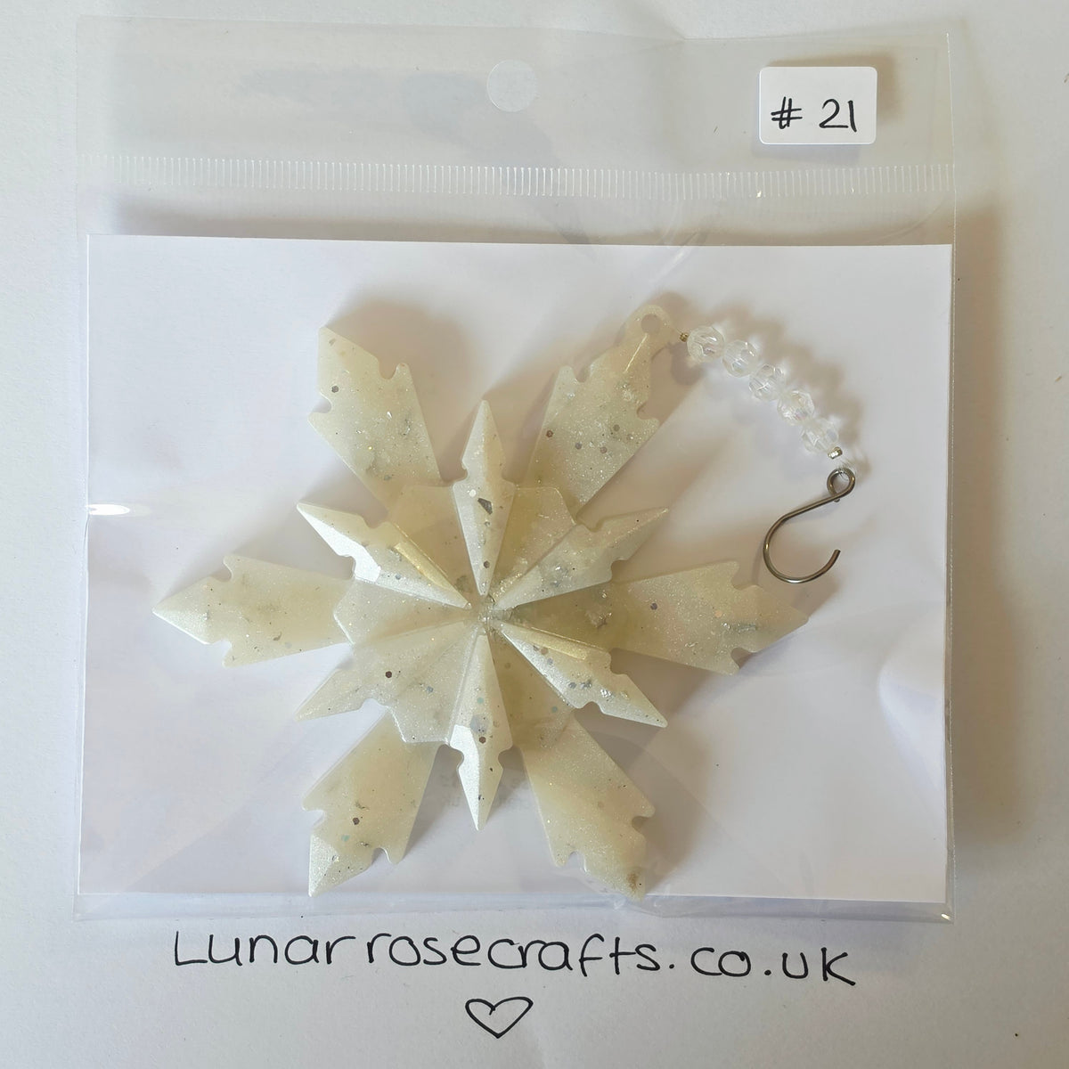A handmade snowflake-shaped hangie made of translucent milky white resin with tiny dark flecks, packaged on a white backing card inside a clear plastic bag. A small white label at the top right reads '# 21', and the card shows 'Lunarrosecrafts.co.uk'