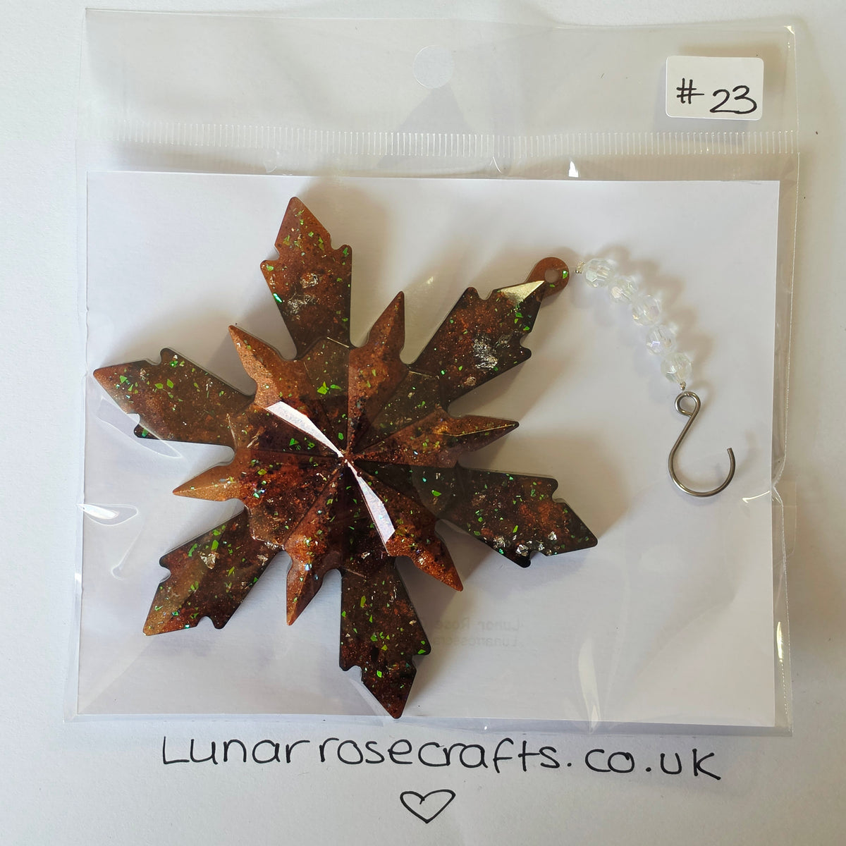 A handmade star-shaped hanging ornament by Lunar Rose Crafts, packaged in a clear plastic bag on a white backing card. It features a metallic bronze-brown glittery finish with green and orange flecks, a white center, and a small white bead chain with a silver hook.