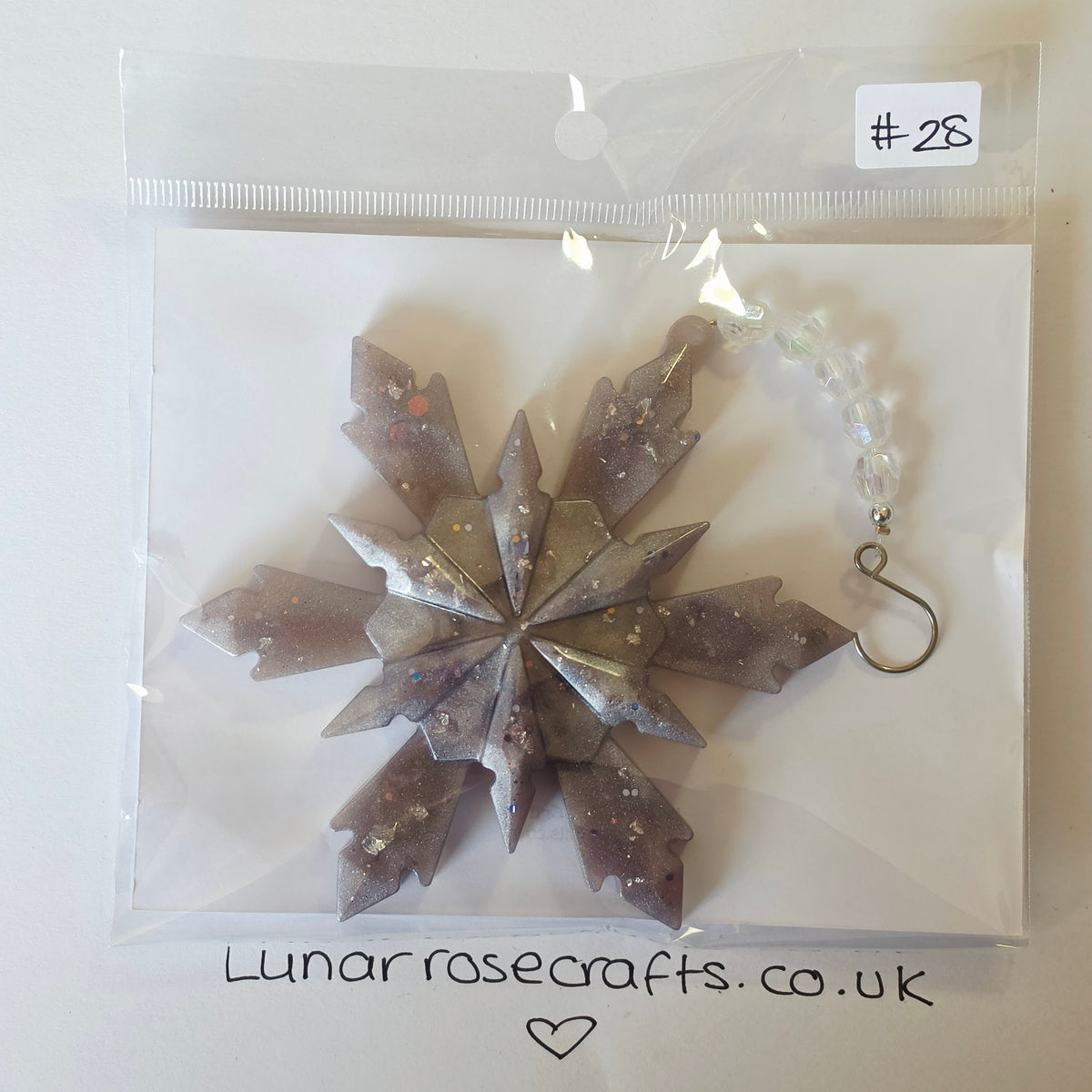 A handmade snowflake ornament in a clear plastic bag with a white insert card. The ornament is made of layered translucent gray and purple resin with embedded glitter, featuring a silver metal hook at the top.