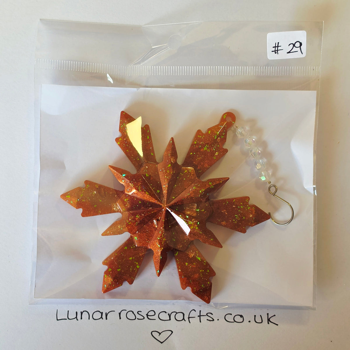 A glittery copper-orange snowflake hanging decoration with a silver metal hanging hook and a clear beaded loop, packaged in a clear plastic zip bag on a white backing card. A handwritten price sticker '#29' is visible in the top right, and 'Lunarrosecrafts.co.uk'